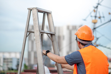 Young Asian maintenance worker with orange safety helmet and vest carrying aluminium step ladder at construction site. Civil engineering, Architecture builder and building service concepts