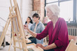 © Seventyfour - Side view portrait of art students sitting in row and painting at easels in art studio, focus on smiling  senior woman enjoying work copy space
