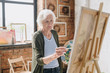 © Seventyfour - Waist up portrait of white haired senior woman holding palette painting pictures at easel in  art studio standing against windows in sunlight, copy space