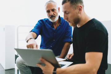 Two bearded men wearing smart casual clothes discussing business strategy of the company by analyzing information in annual company report browsed on a portable computer from the internet.