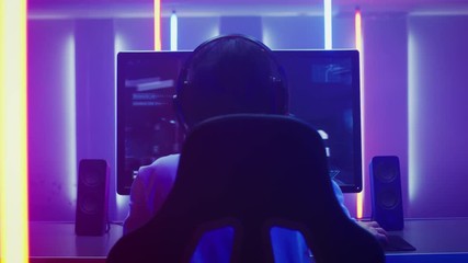 Wall Mural - Back View Shot of the Professional Gamer Playing in First-Person Shooter Online Video Game on His Personal Computer. Room Lit by Neon Lights in Retro Arcade Style.