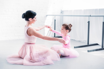 Wall Mural - side view of ballet teacher and little student in pink tutu skirts sitting and holding hands while training in ballet school