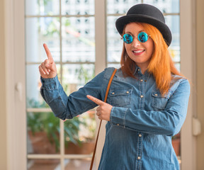 Canvas Print - Stylish redhead woman wearing bowler hat and sunglasses smiling and looking at the camera pointing with two hands and fingers to the side.