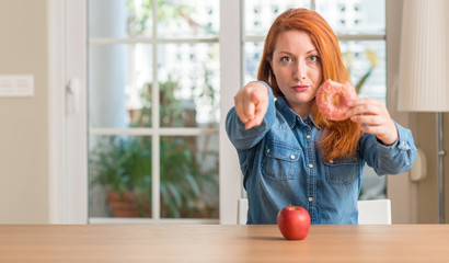 Sticker - Redhead woman chooses between apple and donut pointing with finger to the camera and to you, hand sign, positive and confident gesture from the front