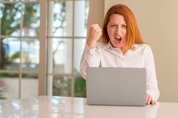 Wall Mural - Redhead woman using computer laptop at home annoyed and frustrated shouting with anger, crazy and yelling with raised hand, anger concept