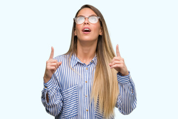 Canvas Print - Beautiful young woman wearing elegant shirt and glasses amazed and surprised looking up and pointing with fingers and raised arms.