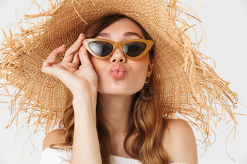 Canvas Print - Portrait of stylish fashionable woman 20s wearing big straw hat and sunglasses giving kiss at camera, isolated over white background