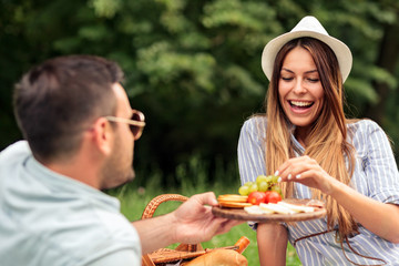 Happy young couple having a great time on a picnic in a park. Man offering fruits and snacks on a wooden plate to his girlfriend. Love and tenderness, dating, romance, lifestyle concept