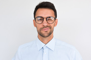 Wall Mural - Portrait of unshaven handsome male frowns, being dissatisfied with something, blink with eye, wears round glasses and blue shirt, isolated over white studio background. People and emotion concept.