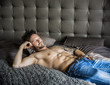 Shirtless sexy male model lying alone on his bed in his bedroom, looking at camera with a seductive attitude