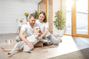 Wall Mural - Young lovely couple in white t-shirts playing with their dog sitting on the floor in the house