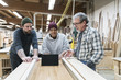 © Mint Images - A group of mixed race carpenters discussing a project at a work station in a large woodworking shop.