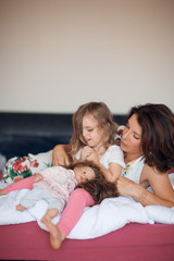 Beautiful happy family scene with mother talking to her little daughter with her doll, lying in bed at home