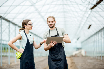Couple of farmers in uniform working with digital tablet standing in the greenhouse with cultivated soil ready for planting