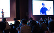 © Right 3 - Presenter with Hand Up Giving Presentation while Audience People Watch in Conference Hall Auditorium. Blurred De-focused Unidentifiable Presenter and Audience. Presentation Screen Video. Technology