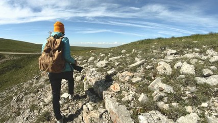 Wall Mural - Kind of a girl going photographer in a hat from behind. A tourist photographer with a backpack and a camera in his hand is walking along a rocky plateau to the precipice to admire the view. Slow