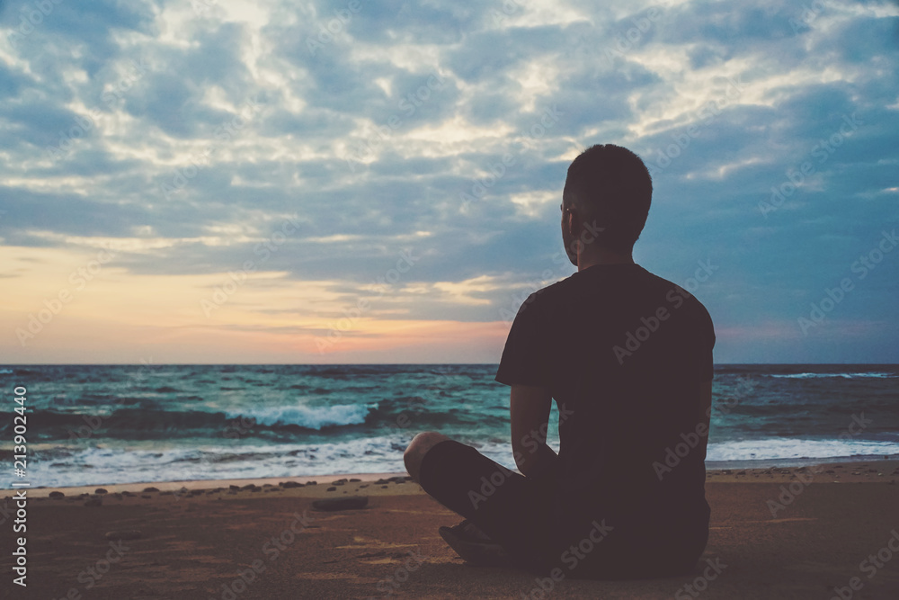 Young man meditating on top ocean cliff during sunset. - obrazy, fototapety, plakaty 
