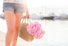 Girl With Straw Bag And Pink Flowers Hydrangea. Traveler Has Vacation In Sea Resort Near Yacht Port At Sunset With Light Background.
