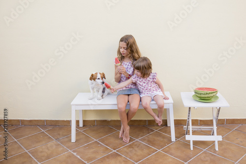 Fotografiet Two beautiful sister kids eating a watermelon ice cream with their cute dog