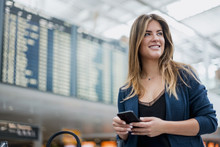 Smiling Young Woman With Cell Phone At Departure Board Looking Around