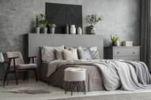 Stylish Bedroom Interior With Bed With Blankets And Pillows, An Armchair, A Footstool, A Drawer Cabinet And A Black Painting On The Wall. Real Photo