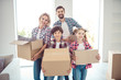 © deagreez - Young happy smiling family four persons wearing casual standing together holding carton boxes with stuff things in light living room moving to new flat
