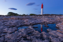 Quiet Evening By A Lighthouse On Rocky Shore. Building Reflected In A Pond. Amazing Colorful Sunset With Only Few Clouds In The Sky.