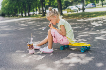 Wall Mural - side view of little child sitting on skateboard and eating dessert by spoon at street