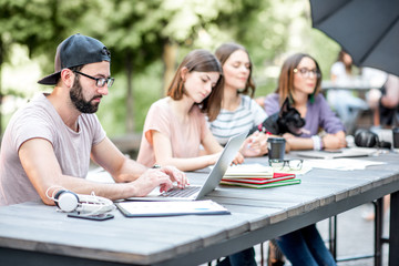 Wall Mural - Young people sitting at the big table working or studying with laptops and documents outdoors in the park