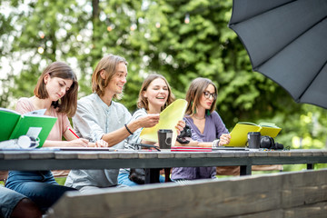 Wall Mural - Young friends dressed casually studying with colorful books sitting in a row at the table outdoors