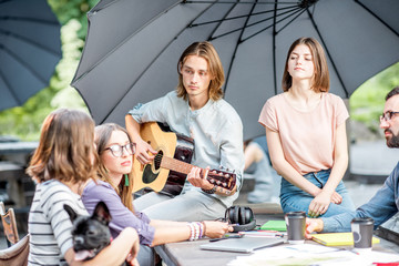 Wall Mural - Young friends having fun together playing a guitar sitting at the table outdoors in the park