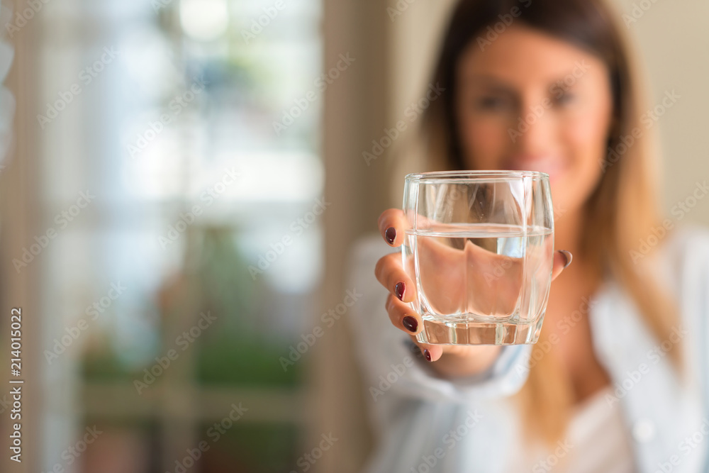 Beautiful young woman smiling while holding a glass of water at home. Lifestyle concept. - obrazy, fototapety, plakaty 
