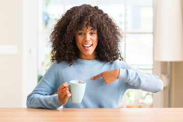 Sticker - African american woman holding a cup of coffee at home with surprise face pointing finger to himself