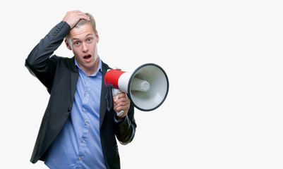 Wall Mural - Young handsome blond man holding a megaphone stressed with hand on head, shocked with shame and surprise face, angry and frustrated. Fear and upset for mistake.