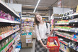 © bodnarphoto - Smiling young woman stands in a supermarket with a shopping cart in her hands and looks at the camera. Portrait of a happy stylish woman doing shopping in a supermarket.