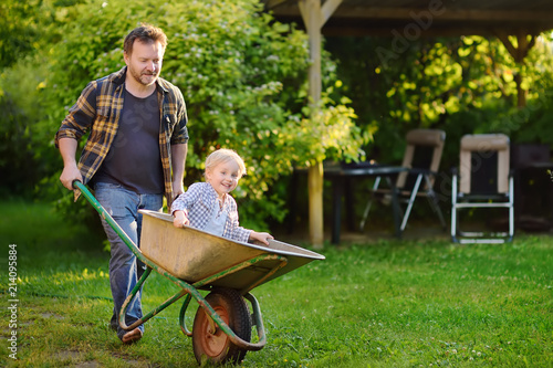 Fotomural Happy little boy having fun in a wheelbarrow pushing by dad in domestic garden