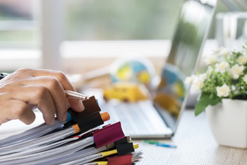 Wall Mural - Businessman hands holding pen for working in Stacks of paper files searching information business report papers and piles of unfinished documents achieves on laptop computer desk in modern office