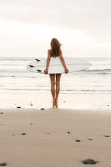 Surf girl with long hair go to surfing. Young surfer woman holding blank white short surfboard on a beach at sunset or sunrise. Bali island, Indonesia. Outdoor Active Lifestyle. It's time for surfing!