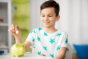 Wall Mural - money, finances, childhood and people concept - smiling little boy putting coin into piggy bank at home