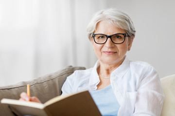 Canvas Print - age, leisure and people concept - happy senior woman in glasses writing to notebook or diary at home