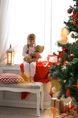 Wall Mural - Cute little child with toy rabbit sitting on windowsill near Christmas tree at home