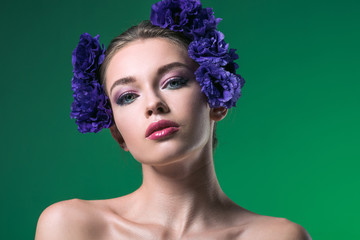 close-up portrait of attractive young woman with eustoma flowers on head looking at camera isolated on green