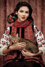 Three Quarter Isolated Portrait Of A Young Slavic Woman In Ethnic Costume, Wearing Embroidered Blouse, Floral Head Scarf, Red Bead Necklace, Holding A Cat On Her Lap, Posing On Dark Brown Background.