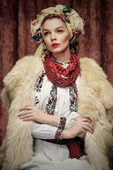 Three quarter isolated studio portrait of a young beautiful Slavic woman in ethnic costume, wearing embroidered blouse, fur coat, floral head scarf, red bead necklace, posing on dark brown background.