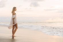 Hipster Trendy Woman In Casual Summer Dress Walking Barefoot By The Waterline And Look To The Waves. Sporty Lady On Sea Sand Beach Sunset Or Ocean Sunrise. Travel, Active, Yoga Lifestyle Concept.