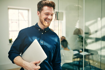 Laughing young businessman walking with his laptop at work