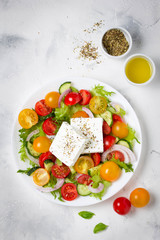 Canvas Print - Greek salad with colorful cherry tomatoes red and yellow, cucumber, onion, lettuce and large piece of feta cheese with herbs. In white plate on light background