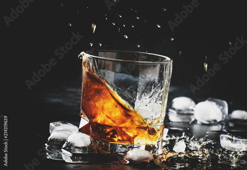 Cuadros en Lienzo Whiskey with ice, splashes out of glass, dark background, toned image, selective