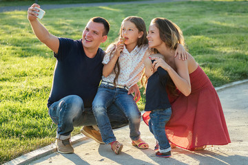 happy family is resting in the park in summer . beautiful mother (woman) , handsome father(man) with a little girl daughter ( schoolgirl ) and baby boy son photographed selfie on phone