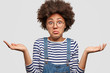 Clueless African American female with curly bushy hair, shrugs shoulders, looks doubtfully as faces dilemma, wears striped sweater and jean overalls, stands against white background. People and doubt
